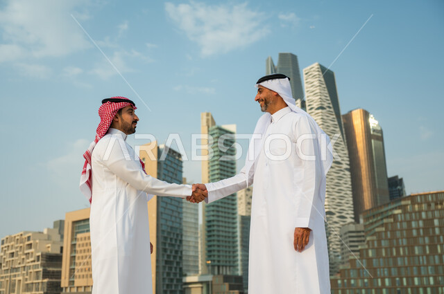 تكوين معارف وتوطيد العلاقات الاجتماعية, إيماءات تدل على السعادة بنجاح الصفقة, اتفاقات عمل بين الشركات الإعمارية, إجراء صفقات عمل بين رجال الأعمال, رجلان عربيان سعوديان خليجيان يرتديان الزي التقليدي يقومان بمصافحة بعضهما, خلفية أبراج مركز عبد الله المالي