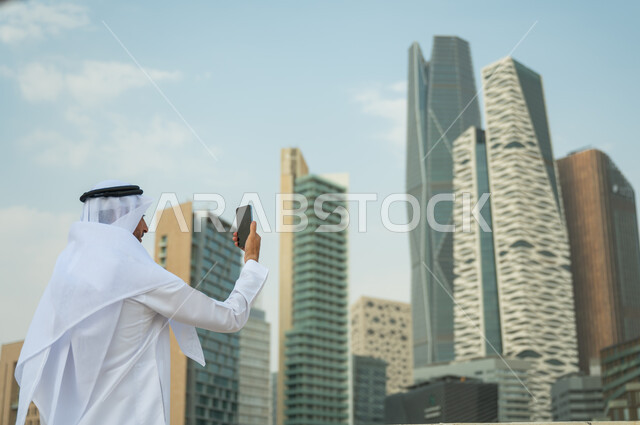 التواصل مع الأصدقاء وإجراء مكالمات فيديو، الأبراج وناطحات السحاب في الرياض في وضح النهار، صورة من الخلف لرجل عربي خليجي سعودي يرتدي الغترة و العقال يقوم بالتقاط صور سيلفي تذكارية باستخدام الهاتف المحمول، خلفية مركز الملك عبد الله المالي