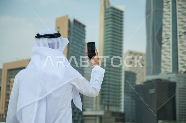 الأبراج وناطحات السحاب في الرياض في وضح النهار، التواصل مع الأصدقاء وإجراء مكالمات فيديو، صورة مقربة من الخلف لرجل عربي خليجي سعودي يرتدي الغترة و العقال يقوم بالتقاط صور سيلفي تذكارية باستخدام الهاتف المحمول، خلفية مركز الملك عبد الله المالي