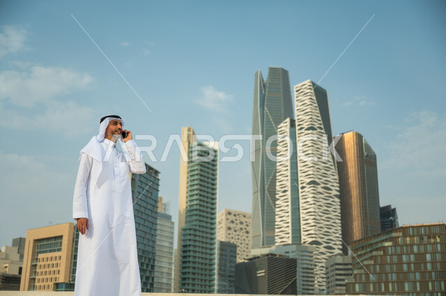 القيام بمكالمة عمل باستخدام الهاتف المحمول, الأبراج المالية في الرياض, التحدث مع الأصدقاء والأهل عبر الجوال، التطور العمراني في الرياض، رجل عربي خليجي سعودي مبتسم يرتدي الغترة والعقال يقوم بإجراء مكالمة صوتية، خلفية مركز الملك عبد الله المالي