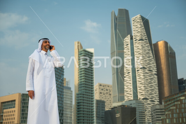 القيام بمكالمة عمل باستخدام الهاتف المحمول, الأبراج المالية في الرياض, التحدث مع الأصدقاء والأهل عبر الجوال، التطور العمراني في الرياض، رجل عربي خليجي سعودي مبتسم يرتدي الغترة والعقال يقوم بإجراء مكالمة صوتية، خلفية مركز الملك عبد الله المالي