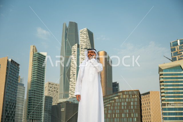 الأبراج المالية في الرياض, القيام بمكالمة عمل باستخدام الهاتف المحمول, التحدث مع الأصدقاء والأهل عبر الجوال، التطور العمراني في الرياض، رجل عربي خليجي سعودي مبتسم يرتدي الغترة والعقال يقوم بإجراء مكالمة صوتية، خلفية مركز الملك عبد الله المالي