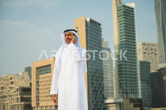 الأبراج المالية في الرياض, القيام بمكالمة عمل باستخدام الهاتف المحمول, التحدث مع الأصدقاء والأهل عبر الجوال، التطور العمراني في الرياض، رجل عربي خليجي سعودي مبتسم يرتدي الغترة والعقال يقوم بإجراء مكالمة صوتية، خلفية مركز الملك عبد الله المالي