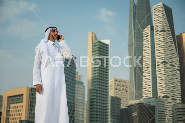Financial towers in Riyadh, making a business call using a mobile phone, talking to friends and family via mobile phone, urban development in Riyadh, a smiling Saudi Arabian Gulf man wearing a ghutra and headband making a voice call, the background of Kin