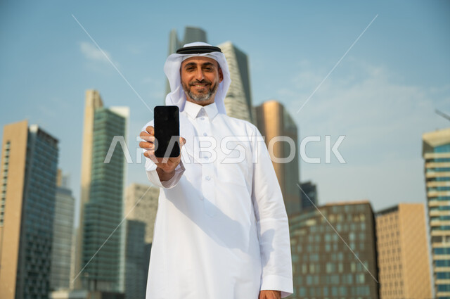 البرامج والتطبيقات العقارية والمالية في السعودية, صورة مقربة لرجل عربي سعودي خليجي مبتسم يرتدي الغترة والثوب التقليدي ويحمل بيده الهاتف المحمول بشاشة سوداء فارغة, عرض مواقع وتطبيقات الجوال, الأبراج المالية في الرياض, خلفية مركز الملك عبد الله المالي