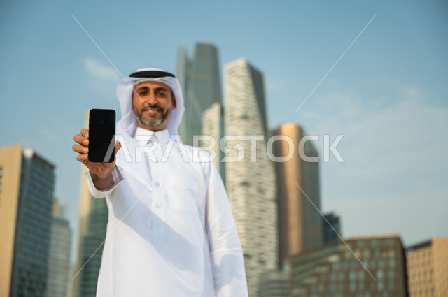 البرامج والتطبيقات العقارية والمالية في السعودية, صورة مقربة لرجل عربي سعودي خليجي مبتسم يرتدي الغترة والثوب التقليدي ويحمل بيده الهاتف المحمول بشاشة سوداء فارغة, عرض مواقع وتطبيقات الجوال, الأبراج المالية في الرياض, خلفية مركز الملك عبد الله المالي