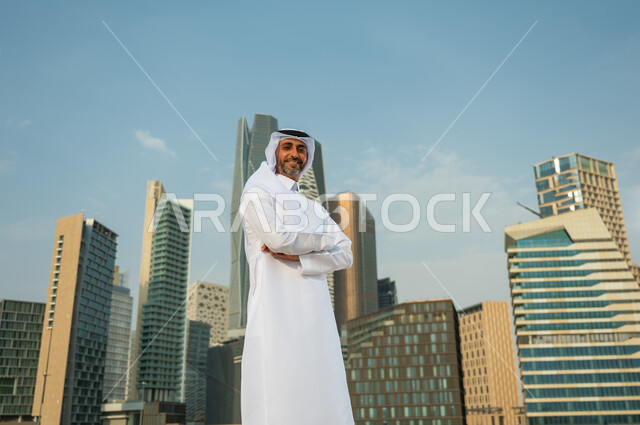 The pride of the people of the Kingdom in its economic development, financial and economic skyscrapers in Riyadh, a Saudi Arabian Gulf man wearing the ghutra and the Saudi thobe, standing with crossed hands with facial gestures indicating happiness, the g