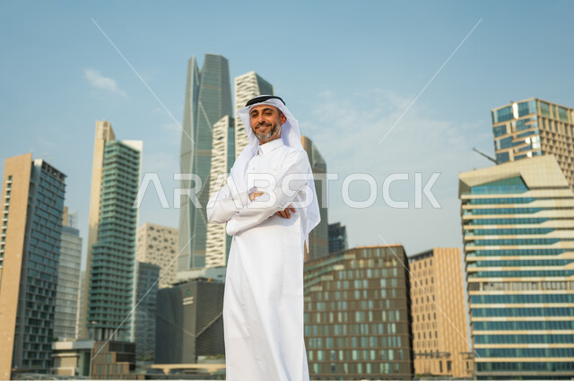 افتخار أبناء المملكة بتطورها الاقتصادي, ناطحات سحاب مالية اقتصادية في الرياض، رجل عربي خليجي سعودي يرتدي الغترة والثوب السعودي يقف بأيدي متقاطعة بإيماءات وجه تدل على السعادة، االنمو والتطور العمراني السعودية، خلفية مركز الملك عبد الله المالي (كافد)