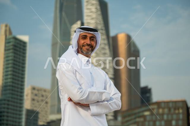 ناطحات سحاب مالية اقتصادية في الرياض، افتخار أبناء المملكة بتطورها الاقتصادي, صورة مقربة لرجل عربي خليجي سعودي يرتدي الغترة والثوب السعودي يقف بأيدي متقاطعة بإيماءات وجه تدل على السعادة، االنمو والتطور العمراني السعودية، خلفية مركز الملك عبد الله المالي