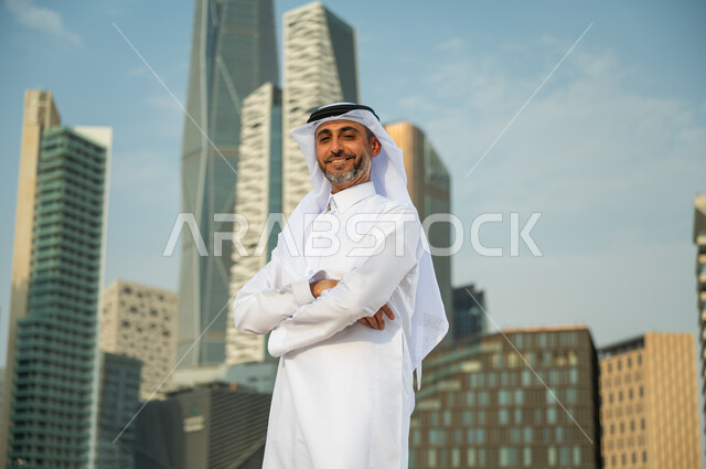 ناطحات سحاب مالية اقتصادية في الرياض، افتخار أبناء المملكة بتطورها الاقتصادي, صورة مقربة لرجل عربي خليجي سعودي يرتدي الغترة والثوب السعودي يقف بأيدي متقاطعة بإيماءات وجه تدل على السعادة، االنمو والتطور العمراني السعودية، خلفية مركز الملك عبد الله المالي