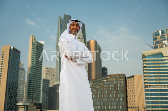 النمو والتطور العمراني السعودية، افتخار أبناء المملكة بتطورها الاقتصادي, ناطحات سحاب مالية اقتصادية في الرياض، رجل عربي خليجي سعودي يرتدي الغترة والثوب السعودي يقف بأيدي متقاطعة بإيماءات وجه تدل على السعادة، خلفية مركز الملك عبد الله المالي (كافد)