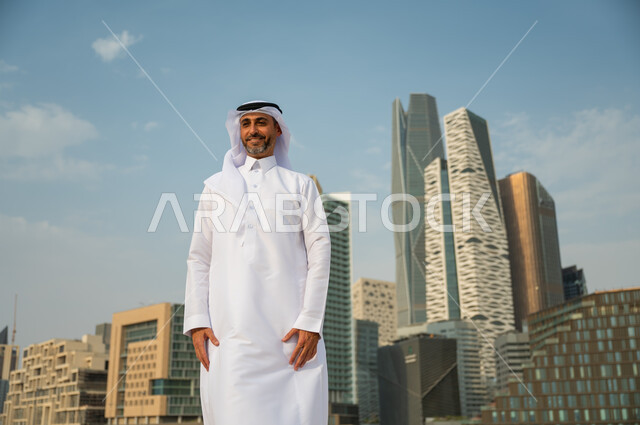 إيماءات الافتخار و الاعتزاز بتطور المملكة, معالم وأماكن سياحية في الرياض, الأبراج المالية الاقتصادية في الرياض، رجل عربي خليجي سعودي مبتسم يرتدي الغترة والعقال يقف باستقامة وينظر الى الكاميرا، االنمو العمراني السعودية، خلفية مركز الملك عبد الله المالي