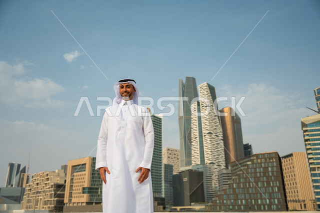 إيماءات الافتخار و الاعتزاز بتطور المملكة, معالم وأماكن سياحية في الرياض, الأبراج المالية الاقتصادية في الرياض، رجل عربي خليجي سعودي مبتسم يرتدي الغترة والعقال يقف باستقامة وينظر الى الكاميرا، االنمو العمراني السعودية، خلفية مركز الملك عبد الله المالي