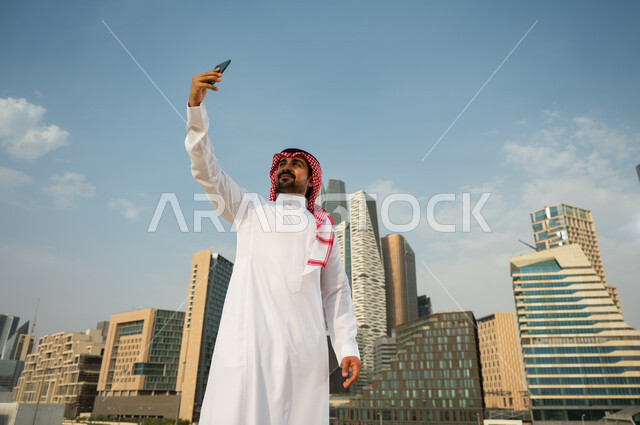 استخدام الجوال لتسجيل اللحظات السعيدة, أخذ لقطات تذكارية مع المعالم السياحية، الأبراج وناطحات السحاب في الرياض في وضح النهار، رجل عربي خليجي سعودي يرتدي الشماغ و العقال يقوم بالتقاط صور سيلفي باستخدام الهاتف المحمول، خلفية أبراج مركز الملك عبد الله المالي