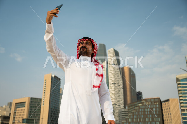 أخذ لقطات تذكارية مع الأبراج وناطحات السحاب في الرياض في وضح النهار، استخدام الجوال لتسجيل اللحظات السعيدة, صورة مقربة لرجل عربي خليجي سعودي يرتدي الشماغ و العقال يقوم بالتقاط صور سيلفي باستخدام الهاتف المحمول، خلفية أبراج مركز الملك عبد الله المالي