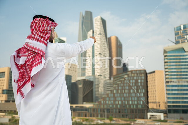 الفن الهندسي المعماري المتميز في الرياض, الإشارة الى ناطحات السحاب  في الرياض، صورة من الخلف لرجل عربي خليجي سعودي يرتدي الشماغ والثوب التقليدي و يشير بيده الى الأبراج المالية في كافد ، االنمو والتطور العمراني السعودية، خلفية مركز الملك عبد الله المالي