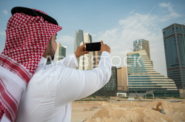 مشاركة التطور الحضاري والعمراني السعودي، نشر معالم الرياض عبر الانترنت, صورة مقربة من الخلف لشاب عربي خليجي سعودي يرتدي الشماغ والزي السعودي يحمل بيده الجوال بشاشة سوداء فارغة، التقاط صور تذكارية لأبراج مركز الملك عبد الله المالي بإستخدام الهاتف المحمول