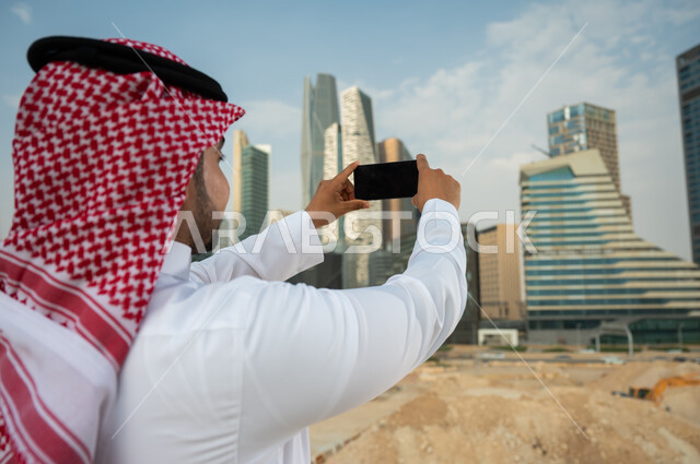 مشاركة التطور الحضاري والعمراني السعودي، نشر معالم الرياض عبر الانترنت, صورة مقربة من الخلف لشاب عربي خليجي سعودي يرتدي الشماغ والزي السعودي يحمل بيده الجوال بشاشة سوداء فارغة، التقاط صور تذكارية لأبراج مركز الملك عبد الله المالي بإستخدام الهاتف المحمول