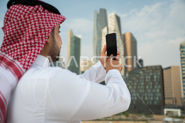 نشر معالم الرياض عبر الانترنت, مشاركة التطور الحضاري والعمراني السعودي، صورة مقربة من الخلف لشاب عربي خليجي سعودي يرتدي الشماغ والزي السعودي يحمل بيده الجوال بشاشة سوداء فارغة، التقاط صور تذكارية لأبراج مركز الملك عبد الله المالي بإستخدام الهاتف المحمول