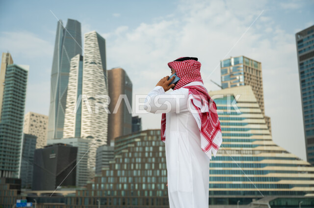 ناطحات السحاب المالية في الرياض, إجراء مكالمة عمل عبر الجوال, دردشات مع الأهل والأصدقاء عبر الموبايل، النمو العمراني في الرياض، صورة من الجانب لرجل عربي خليجي سعودي مبتسم يرتدي الشماغ والعقال يقوم بإجراء مكالمة صوتية، خلفية مركز الملك عبد الله المالي