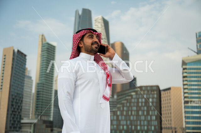 النمو العمراني في الرياض، إجراء مكالمة عمل عبر الجوال, ناطحات السحاب المالية في الرياض, دردشات مع الأهل والأصدقاء عبر الموبايل، صورة من الجانب لرجل عربي خليجي سعودي مبتسم يرتدي الشماغ والعقال يقوم بإجراء مكالمة صوتية، خلفية مركز الملك عبد الله المالي