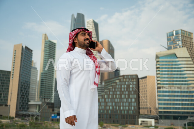 النمو العمراني في الرياض، إجراء مكالمة عمل عبر الجوال, ناطحات السحاب المالية في الرياض, دردشات مع الأهل والأصدقاء عبر الموبايل، صورة من الجانب لرجل عربي خليجي سعودي مبتسم يرتدي الشماغ والعقال يقوم بإجراء مكالمة صوتية، خلفية مركز الملك عبد الله المالي