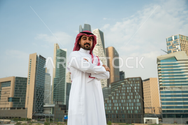 التباهي بتطور المملكة الاقتصادي, ناطحات سحاب مالية اقتصادية في الرياض، صورة مقربة لرجل عربي خليجي سعودي يرتدي الشماغ والثوب السعودي يقف بأيدي متقاطعة بإيماءات وجه تدل على الافتخار، االنمو والتطور العمراني السعودية، خلفية مركز الملك عبد الله المالي
