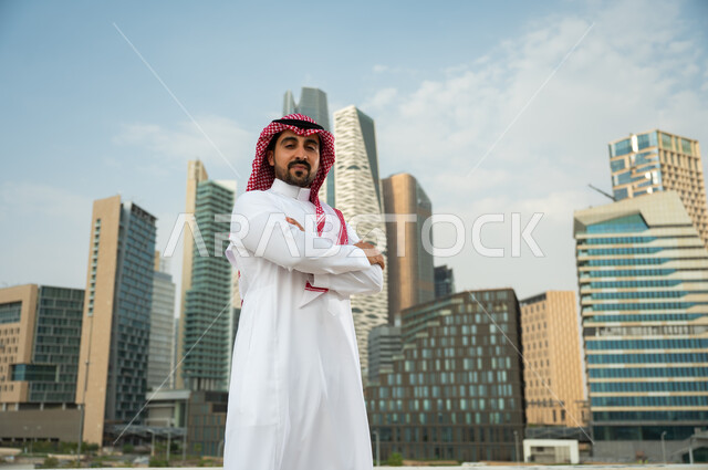 النمو والتطور العمراني السعودية، التباهي بتطور المملكة الاقتصادي, ناطحات سحاب مالية اقتصادية في الرياض، صورة مقربة لرجل عربي خليجي سعودي يرتدي الشماغ والثوب السعودي يقف بأيدي متقاطعة بإيماءات وجه تدل على الافتخار، خلفية مركز الملك عبد الله المالي