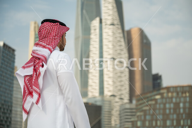 Standing in front of the skyscrapers in Riyadh, the development and distinguished architectural engineering art in Riyadh, a back image of a Saudi Arabian Gulf man wearing the shamagh and traditional dress and standing upright and proud, the Saudi urban d