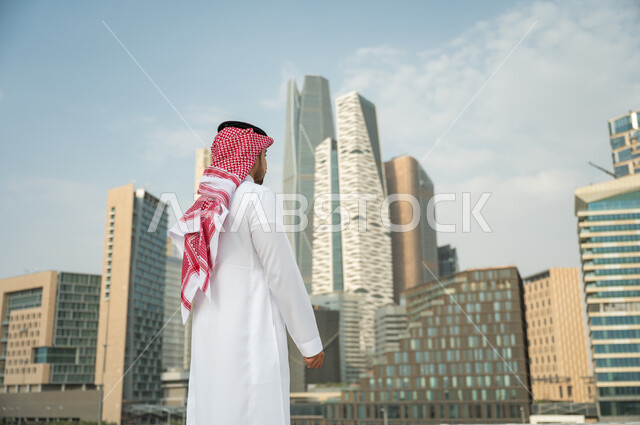 الوقوف أمام ناطحات السحاب  في الرياض، التطور و الفن الهندسي المعماري المتميز في الرياض, صورة من الخلف لرجل عربي خليجي سعودي يرتدي الشماغ والثوب التقليدي و يقف باستقامة وشموخ، التطور العمراني السعودي، خلفية مركز الملك عبد الله المالي
