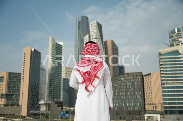 التطور العمراني السعودي، التطور و الفن الهندسي المعماري المتميز في الرياض, الوقوف أمام ناطحات السحاب  في الرياض، صورة من الخلف لرجل عربي خليجي سعودي يرتدي الشماغ والثوب التقليدي و يقف باستقامة وشموخ، خلفية مركز الملك عبد الله المالي