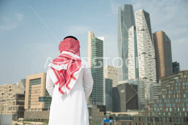 التطور العمراني السعودي، التطور و الفن الهندسي المعماري المتميز في الرياض, الوقوف أمام ناطحات السحاب  في الرياض، صورة من الخلف لرجل عربي خليجي سعودي يرتدي الشماغ والثوب التقليدي و يقف باستقامة وشموخ، خلفية مركز الملك عبد الله المالي
