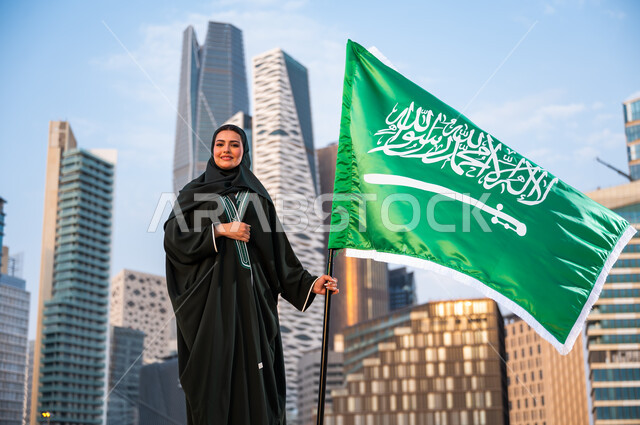 Day of unity and the modern history of the Kingdom, embodying feelings of love and loyalty, celebrating the Saudi National Day, National Day on the 8th of Rabi’ al-Awwal, a veiled Saudi Gulf Arab woman wearing the abaya and holding the Kingdom’s flag with pride and appreciation, Saudi Flag Day March 11, background of the Kafed Towers in Riyadh