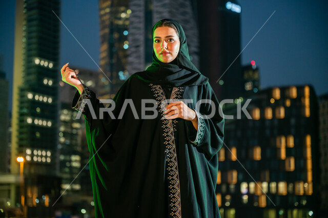 انعكاس الضوء الأخضر على مواطنة عربية سعودية خليجية ترتدي الزي التقليدي بذكرى اليوم الوطني23 سبتمبر, المشاركة في المناسبات الوطنية, النظر إلى الكاميرا بإيماءات الإعجاب بالعباءة المطرزة, يوم العلم السعودي 11 مارس, خلفية الأبراج والمباني في مدينة الرياض