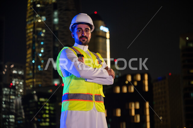 التطور والتقدم العمراني، الوقوف بفخر واعتزاز والنظر إلى الكاميرا بإيماءات أيدي متقاطعة, مفهوم الإنشاءات و الهندسة المعمارية, مهندس سعودي مبتسم يرتدي سترة العمل وخوذة الحماية البيضاء في وقت الليل , خلفية الأبراج وناطحات السحاب المضيئة ليلا في مدينة الرياض