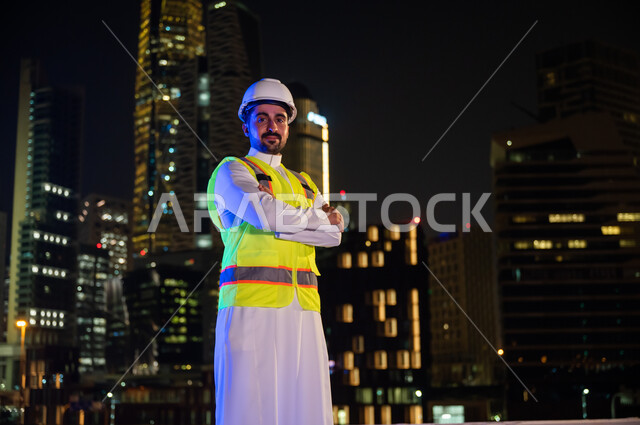 Urban progress and development, standing proudly and looking at the camera with crossed hand gestures, the concept of construction and architecture, a smiling Saudi engineer wearing a work jacket and a white protection helmet at night time, the background