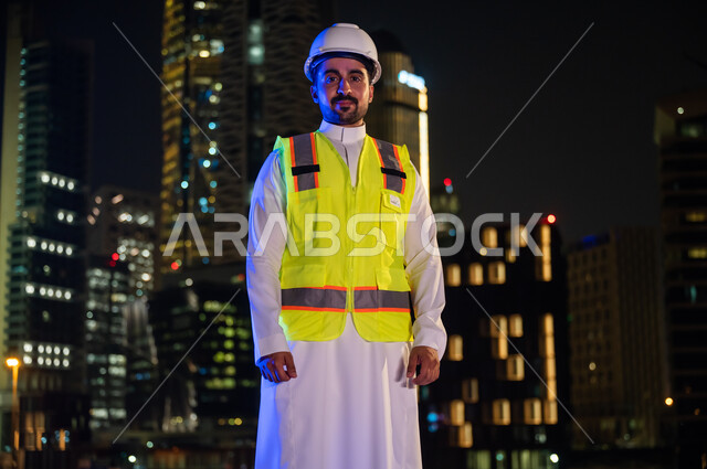 Standing with pride and pride and looking at the camera, interest in urban development with the arms of Saudi engineers, a smiling Saudi Arab Gulf engineer wearing a work jacket and a white protection helmet at night, crossed hands gestures, the backgroun