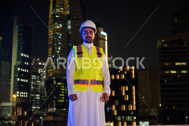 الاهتمام بالتطور العمراني بسواعد المهندسين السعوديين، الوقوف بفخر واعتزاز والنظر إلى الكاميرا, مهندس عربي خليجي سعودي مبتسم يرتدي سترة العمل وخوذة الحماية البيضاء ليلا ,ايماءات أيدي متقاطعة, خلفية الأبراج وناطحات السحاب المضيئة ليلا في مدينة الرياض