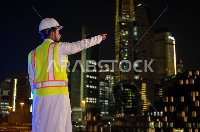Referring to something, the concept of architecture and construction, the progress and development of Saudi buildings at the hands of Saudi engineers, a picture from the back of a Saudi Arab Gulf engineer wearing a work jacket and a white protection helme