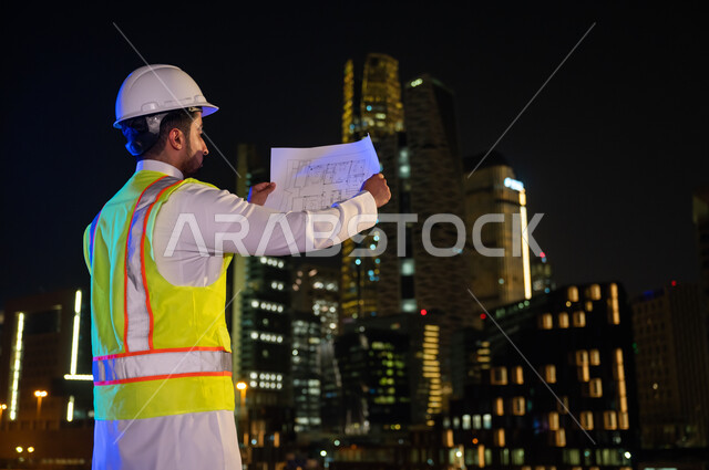 The concept of architecture and construction, the progress and development of Saudi buildings by Saudi engineers, the background of towers and skyscrapers illuminated at night in the city of Riyadh, a close-up from the back of a Saudi Arabian Gulf enginee