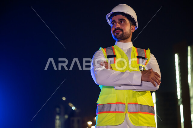 الاهتمام بالتطوير العمراني، مفهوم الإنشاءات و الهندسة المعمارية, الوقوف بفخر واعتزاز والنظر إلى شيء ما, صورة مقرب لمهندس عربي خليجي سعودي مبتسم يرتدي سترة العمل وخوذة الحماية البيضاء في وقت الليل ,ايماءات أيدي متقاطعة
