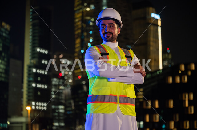 The concept of construction and architecture, standing proudly and looking at something, a smiling Saudi Arabian Gulf engineer wearing a work jacket and a white protection helmet at night time, crossed hands gestures, the background of the towers and skys