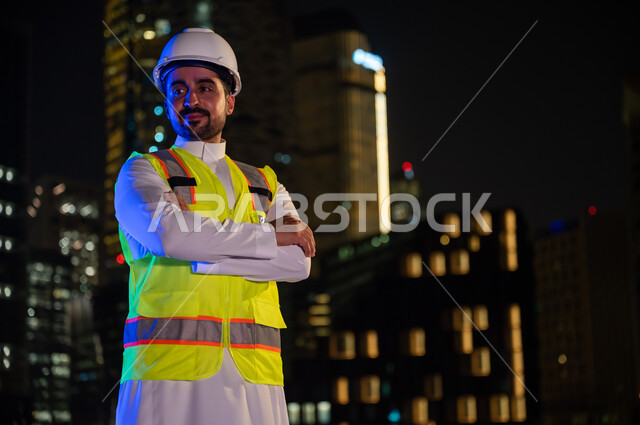 الوقوف بفخر واعتزاز والنظر إلى شيء ما, مفهوم الإنشاءات و الهندسة المعمارية, مهندس عربي خليجي سعودي مبتسم يرتدي سترة العمل وخوذة الحماية البيضاء في وقت الليل ,ايماءات أيدي متقاطعة, خلفية الأبراج وناطحات السحاب المضيئة ليلا في مدينة الرياض .