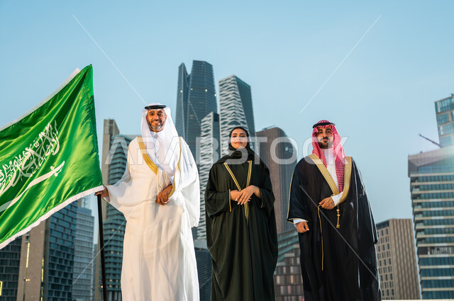 Standing confidently on Flag Day, March 11, wearing traditional folk costumes, celebrating Saudi national occasions, Saudi Gulf Arab citizens celebrating National Day, September 23, looking at the camera with gestures of confidence and pride, background of the economic towers in Riyadh