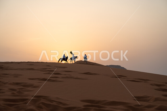 أنشطة وفعاليات ركوب الجياد في الصحراء، ذكرى اليوم الوطني السعودي 8 ربيع الأول، رفع الراية للاحتفال بيوم العلم السعودي 11 مارس، مجموعة رجال عرب خليجيون سعوديون يتجولون على ظهر الاحصنة وقت غروب الشمس، حب وعشق الخيل السعودي الأصيل.