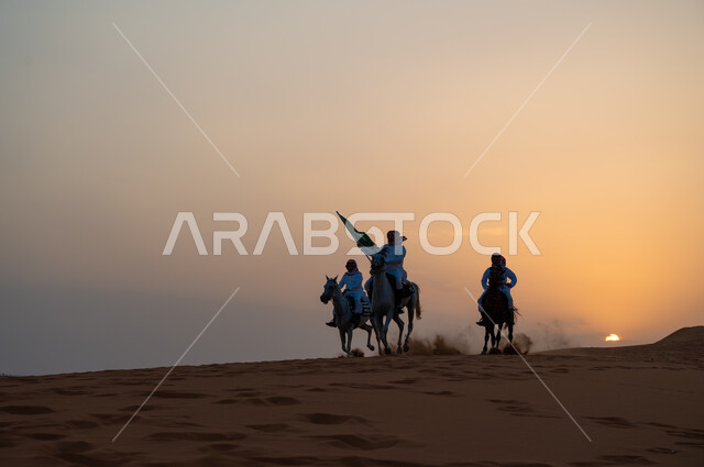 أنشطة وفعاليات ركوب الجياد في الصحراء، ذكرى اليوم الوطني السعودي 8 ربيع الأول، رفع الراية للاحتفال بيوم العلم السعودي 11 مارس، مجموعة رجال عرب خليجيون سعوديون يتجولون على ظهر الاحصنة وقت غروب الشمس، حب وعشق الخيل السعودي الأصيل.