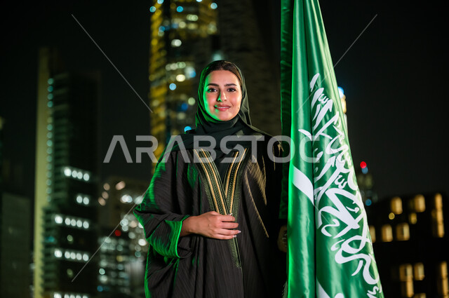 الأبراج والمباني في الرياض ليلا, النظر إلى الكاميرا بإيماءات السعادة, المشاركة بالمناسبات الوطنية, صورة مقربة لإمرأة عربية سعودية خليجية مبتسمة ترتدي العباءة والحجاب تمسك علم المملكة باحترام وتقدير, استذكار اليوم الوطني 23 سبتمبر, يوم العلم 11 مارس