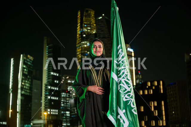 المشاركة بالمناسبات الوطنية, النظر إلى الكاميرا بإيماءات السعادة, إمرأة عربية سعودية خليجية مبتسمة ترتدي العباءة والحجاب تمسك علم المملكة باحترام وتقدير, استذكار اليوم الوطني 23 سبتمبر, يوم العلم 11 مارس, الأبراج والمباني في الرياض ليلا