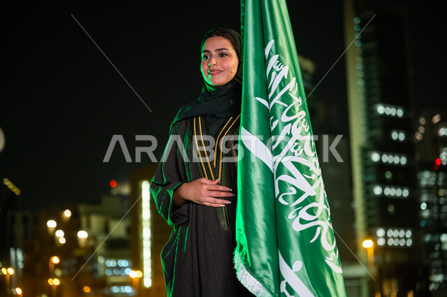 الحب والولاء للدولة بالمشاركة باليوم الوطني السعودي 23 سبتمبر, إمرأة عربية سعودية خليجية محجبة ترتدي العباءة تمسك علم المملكة بفخر وتقدير تنظر إلى شيء ما, يوم العلم السعودي 11 مارس, خلفية المباني المضاءة في وقت الليل في الرياض