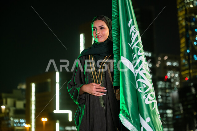 يوم العلم السعودي 11 مارس, الحب والولاء للدولة بالمشاركة باليوم الوطني السعودي 23 سبتمبر,  إمرأة عربية سعودية خليجية محجبة ترتدي العباءة تمسك علم المملكة بفخر وتقدير تنظر إلى شيء ما, خلفية المباني المضاءة في وقت الليل في مدينة الرياض